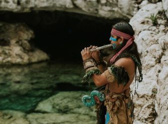 man in traditional maya clothes and face paint standing by water playing instrument