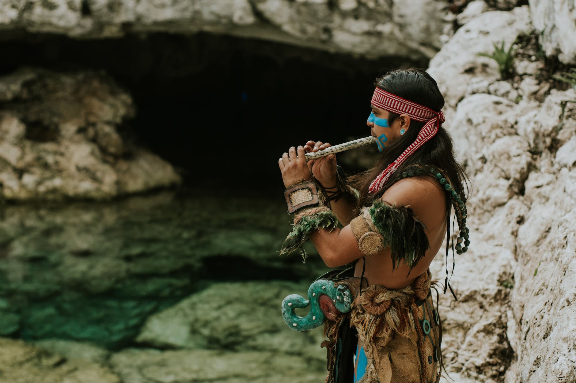 man in traditional maya clothes and face paint standing by water playing instrument